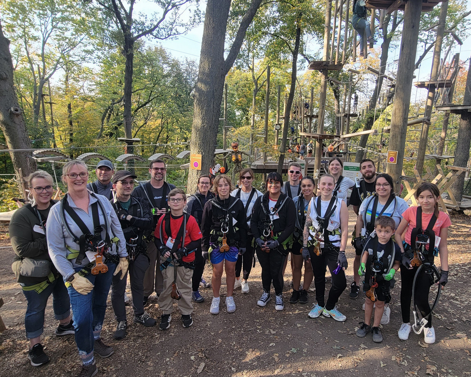 Group of people wearing safety harnesses pose at the base of a forest ropes course with platforms and obstacles in the trees.